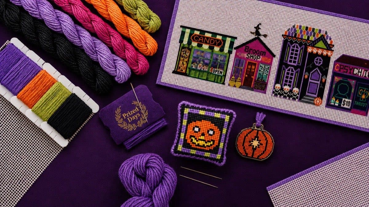 Prized Days needlepoint flat lay — silk threads, needle book, and Halloween village stitchery on plum velvet